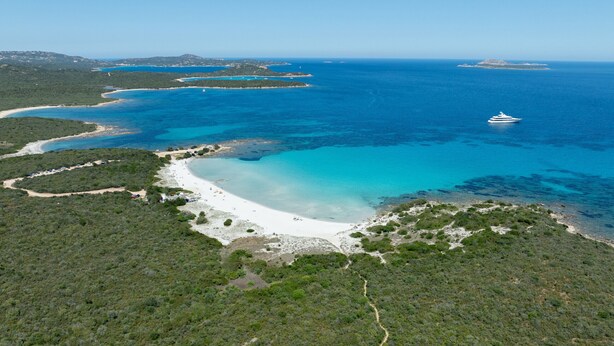 An aerial view of a coastal area with a beach, clear blue waters, and a yacht.