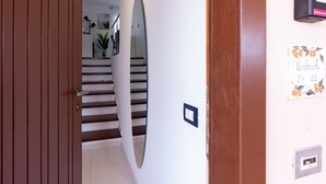 A hallway with a wooden door, a white wall with a mirror, a staircase, and a wall-mounted intercom.