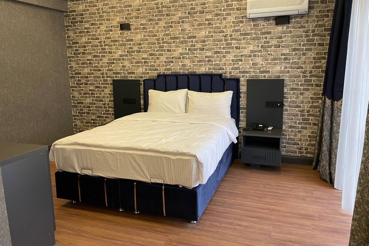 A bedroom with a brick-patterned wall, a bed with a dark headboard, a bedside table with speakers, and a small cabinet.