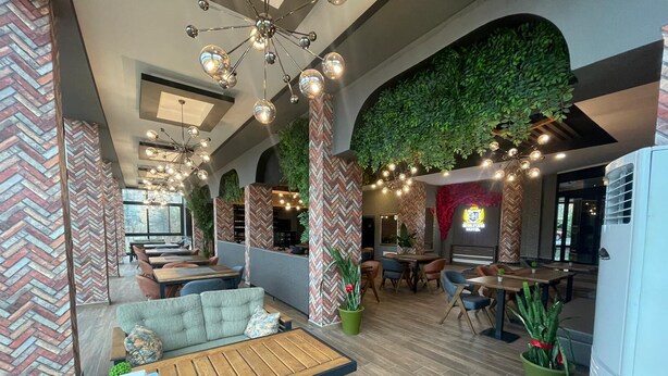 Un restaurant moderne avec de hauts plafonds, des murs en briques apparentes et un décor mural végétal. On y trouve plusieurs espaces de restauration avec des tables et des chaises, ainsi qu’un bar agrémenté d’un néon.