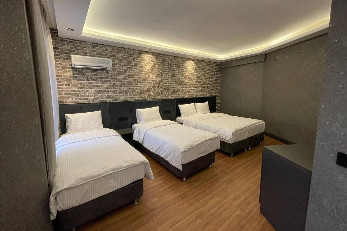 A modern hotel room with three beds, a brick wall, and a recessed ceiling light.
