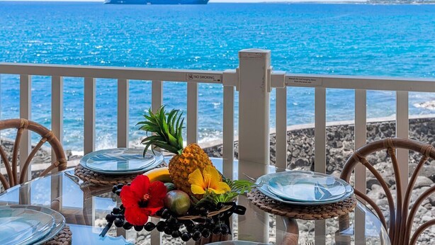 A table with a tropical fruit arrangement and a glass container, overlooking a beach with a cruise ship in the distance.