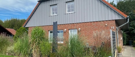 A two-story house with a grey roof and red brick walls, number 15, surrounded by a well-maintained lawn and various plants.