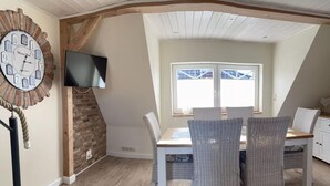 Een eetgedeelte met een witte tafel en rieten stoelen, een klok aan de muur en een televisie boven de tafel bevestigd.