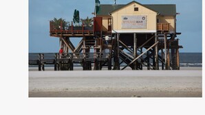 Een strandbar met een bord waarop 'STRANDBAR 54 NORD' staat, op een houten constructie met een groen dak.