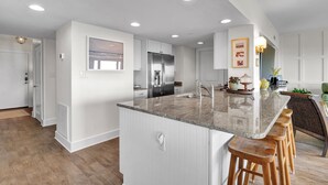 A modern kitchen with white cabinetry, a stainless steel refrigerator, and a granite countertop. - Prime Oceanfront Location: Pool, Balcony & Tennis Court (Wrightsville Beach)