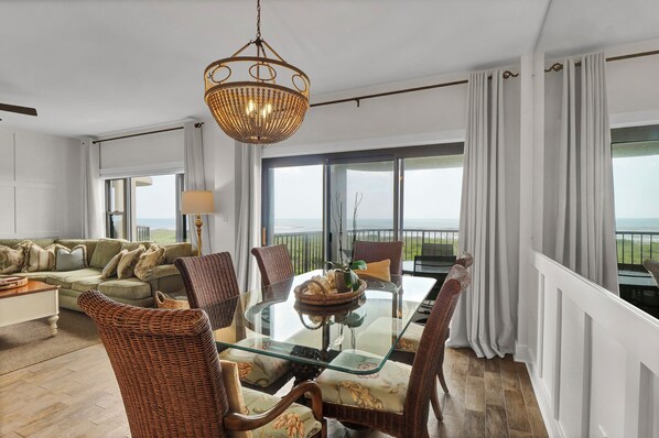 A spacious living room with a glass-top dining table, wicker chairs, a green sofa, and a view of the sea through large windows.