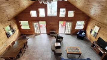 A spacious loft-style room with a high ceiling, large windows, a wooden floor, a sofa, a coffee table, and a TV.
