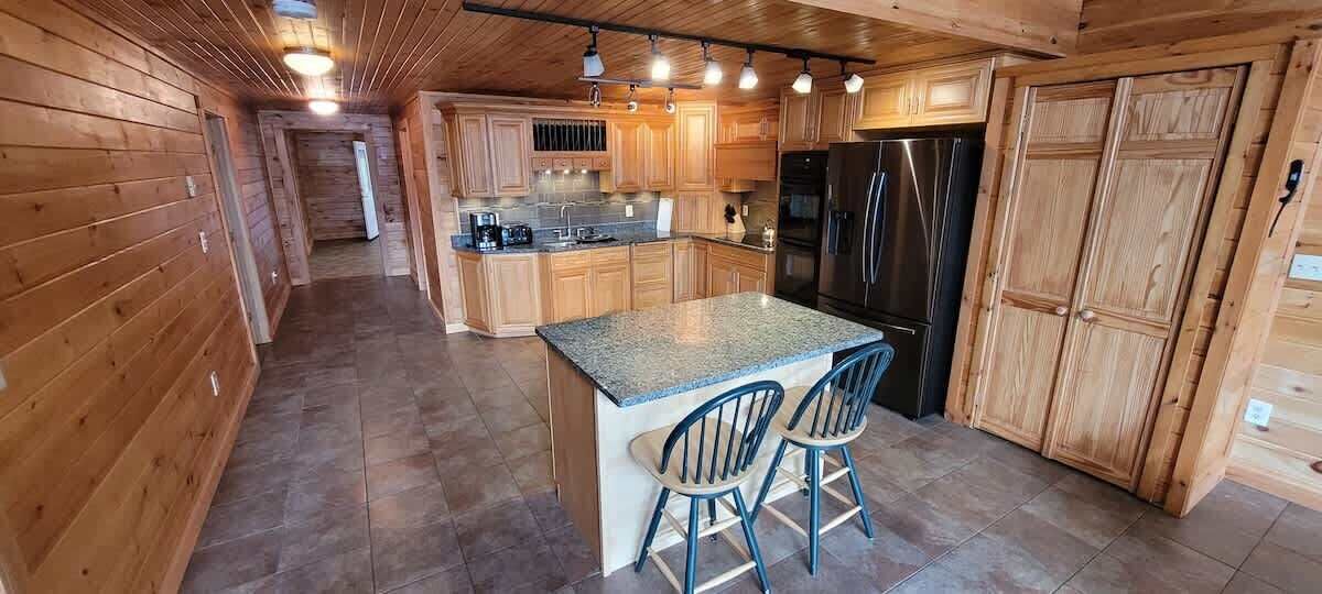 A wooden cabin interior with a kitchen featuring a granite countertop island, two bar stools, and a refrigerator.