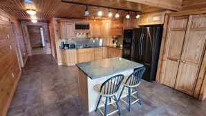 A wooden cabin interior with a kitchen featuring a granite countertop island, two bar stools, and a refrigerator. - Lakeside Luxury: Your Private Waterfront Escape (Lincoln)