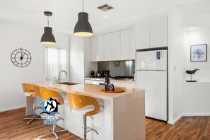 A modern kitchen with white cabinets, a central island with bar stools, pendant lighting, and a refrigerator. - Contemporary Lakeside Stunner – 3 bedrooms & jetty (Encounter Bay)