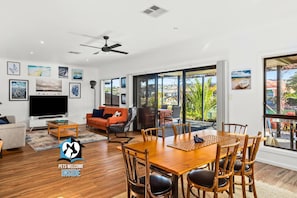 A spacious living room with a large TV, orange sofa, and a dining area with wooden furniture. - Contemporary Lakeside Stunner – 3 bedrooms & jetty (Encounter Bay)