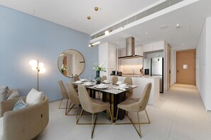 A modern dining area with a round table set for six, surrounded by beige chairs with gold legs. A large mirror hangs on the wall, and a floor lamp is visible. The room has a light blue accent wall and white flooring. - Luxury Living w/ Superb Sea Views in Address JBR (Dubai)
