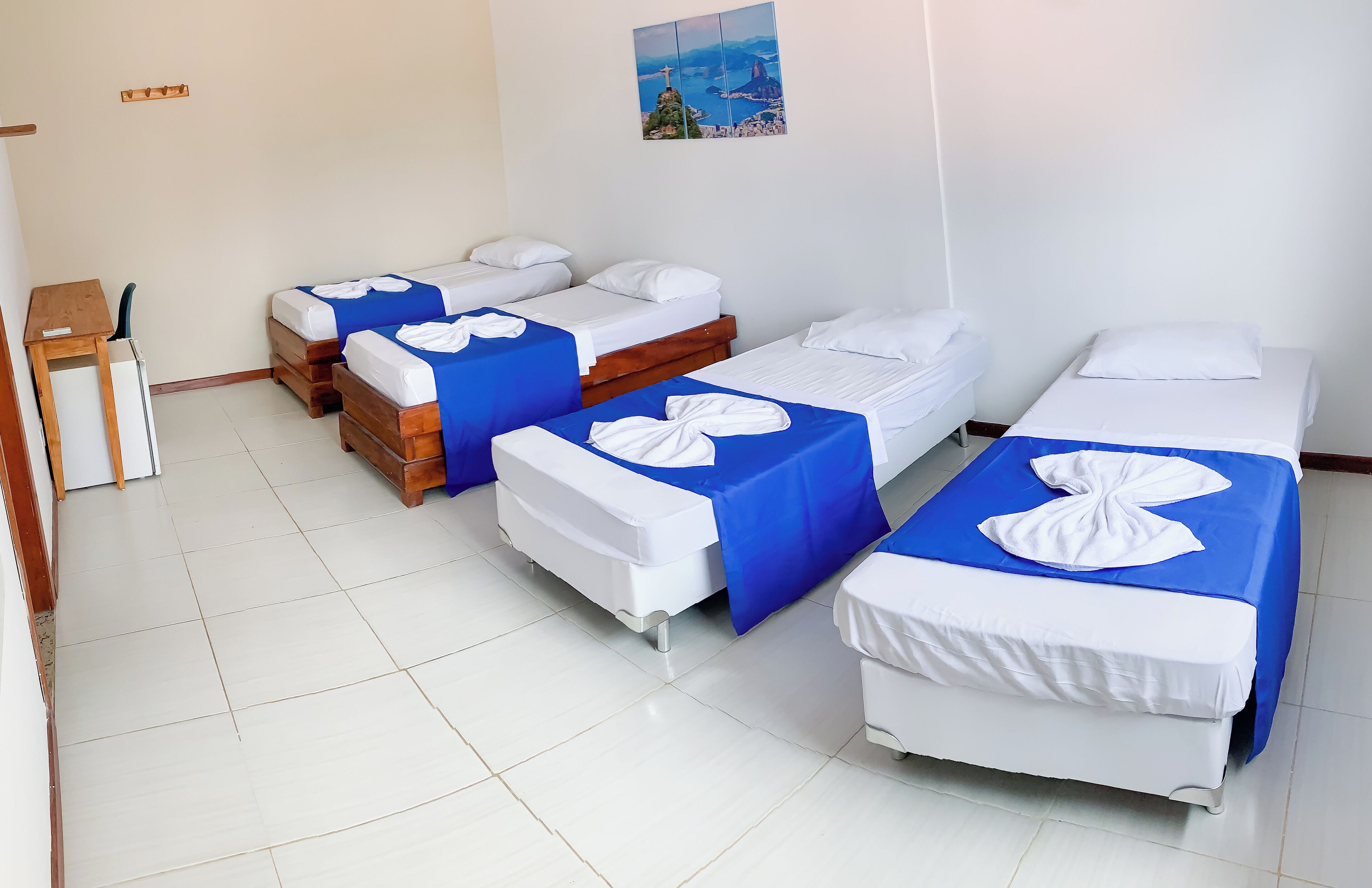 A hotel room with four single beds, each with a blue and white bedspread. There is a small wooden desk with a chair, and a white radiator on the left.