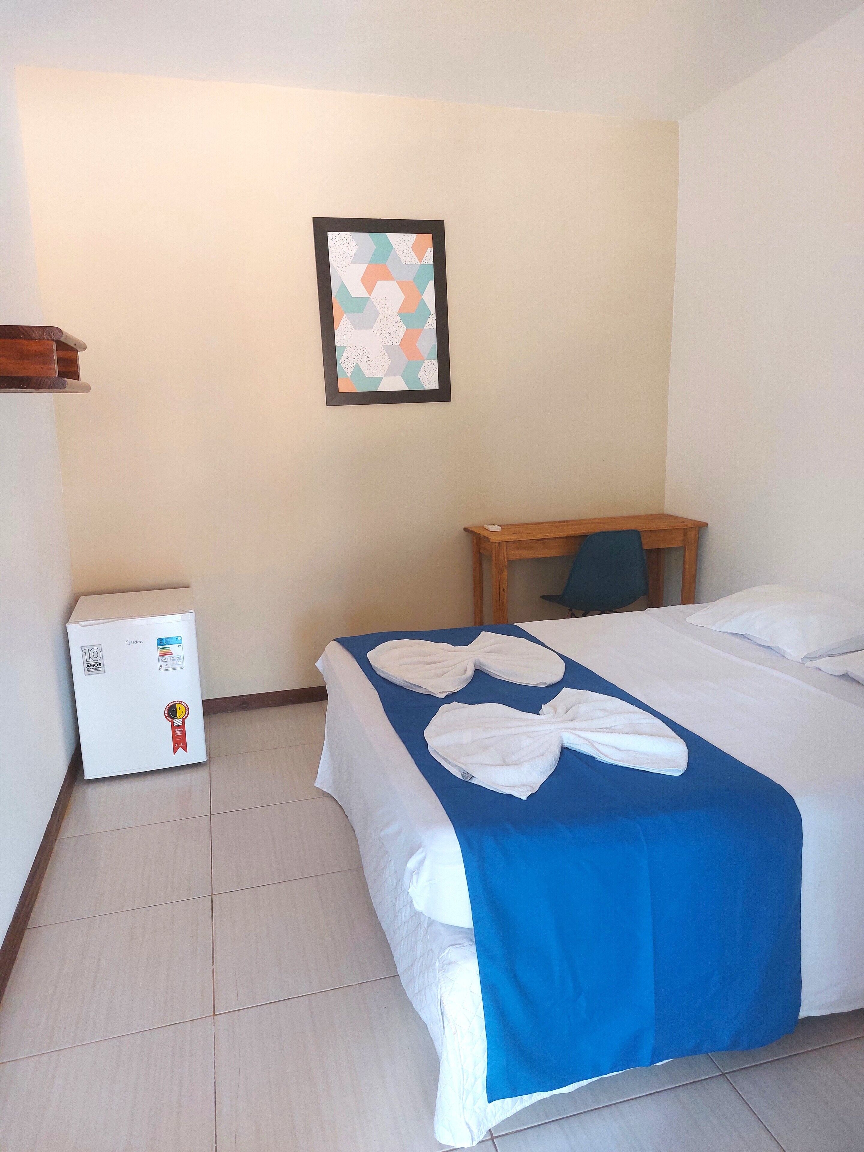 A hotel room with a bed, a small table, a refrigerator, and a framed picture on the wall.