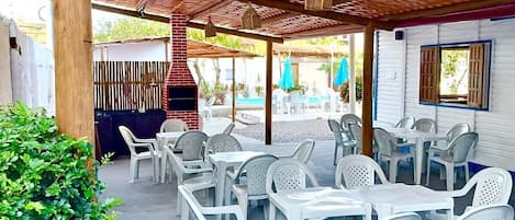 Un patio al aire libre con mesas y sillas de plástico blanco, una pérgola de madera y vistas a una zona de piscina.