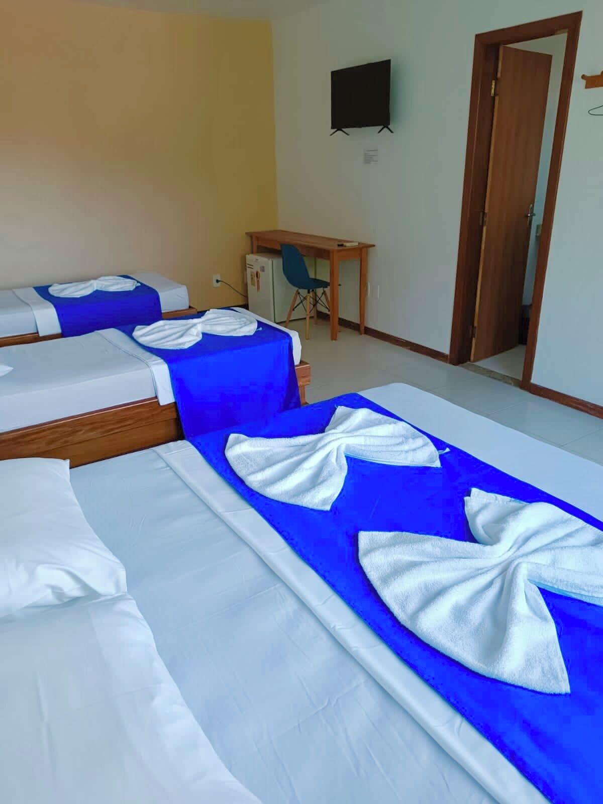 A hotel room with three beds, each with white linens and blue bedspreads. There is a small desk with a chair, a television on the wall, and a door leading to another room.