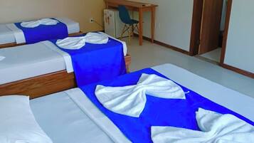 A hotel room with three beds, each with white linens and blue bedspreads. There is a small desk with a chair, a television on the wall, and a door leading to another room.