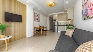 A modern living room with a grey sofa, a flat-screen TV mounted on a wooden panel wall, a dining area with a wooden table and chairs, and a kitchen with a microwave and refrigerator. - Stunning apartment in Costa Adeje with heated pool. (Costa Adeje)