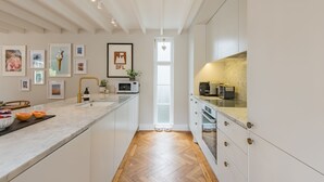 Une cuisine moderne avec des meubles blancs, un plan de travail en marbre et un parquet en bois disposé en chevrons. On peut y voir des tableaux encadrés au mur, une fenêtre décorée d’un motif en forme de cœur et un four à micro-ondes posé sur le plan de travail.