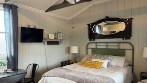 Ein Schlafzimmer mit Bett, Schreibtisch, Stuhl, Fernseher, Spiegel und Deckenventilator.