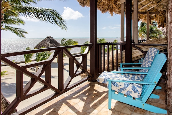 Un balcon donnant sur la plage, avec un toit de chaume, une chaise bleue et une vue sur l’océan.
