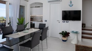 A modern dining area with a glass table and chairs, a kitchen in the background, and a TV mounted on the wall. - Harenae Beach Gataway by Wonderful Italy (Olbia)