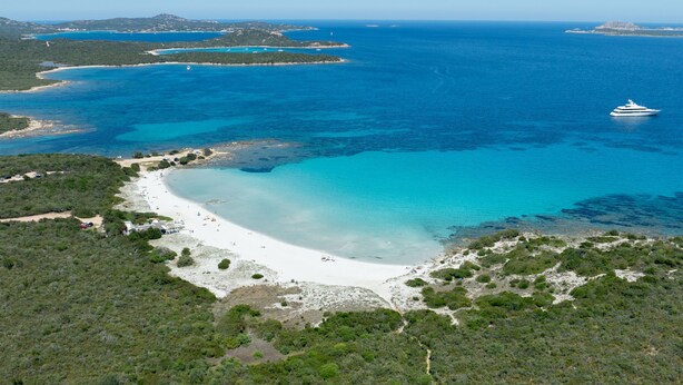 An aerial view of a coastal area with a beach, clear blue waters, and a yacht in the distance. - Harenae Beach Gataway by Wonderful Italy (Olbia)