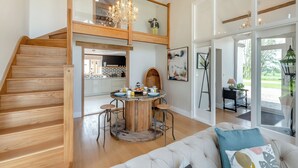 Ein geräumiges Loft mit einer Holztreppe, einem Essbereich mit rundem Tisch und einem großen Fenster mit Blick ins Freie.