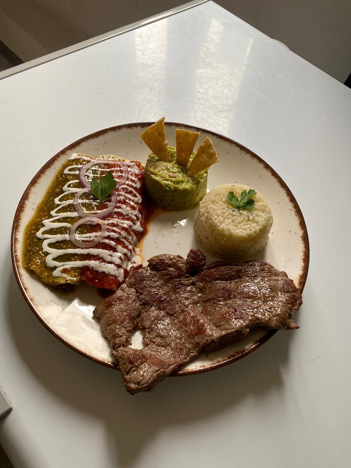 A plate with a steak, rice, guacamole, and a red sauce with onion rings.