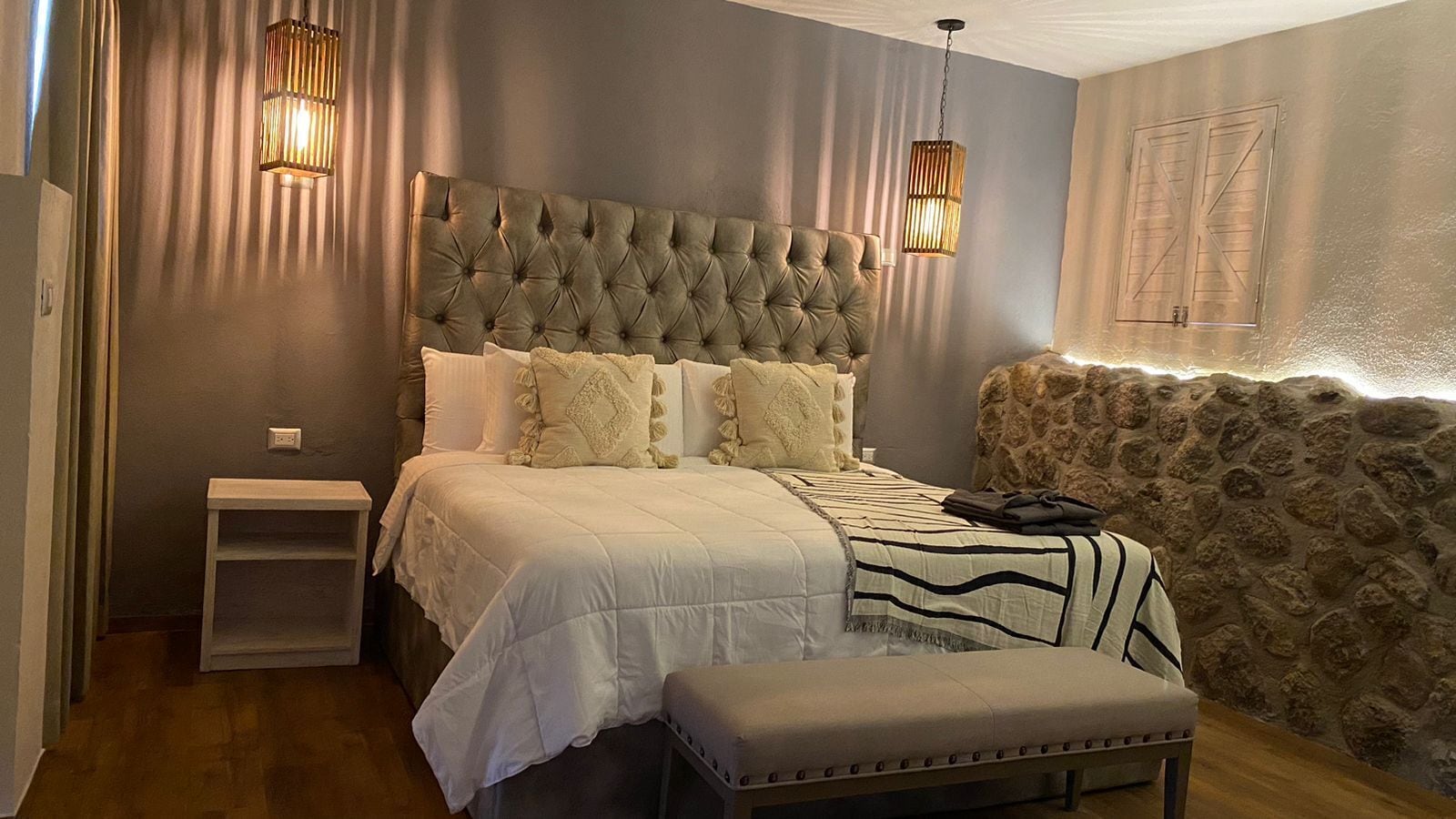 Une chambre à coucher avec une tête de lit capitonnée, une table de chevet, un banc et un mur d’accent en pierre.