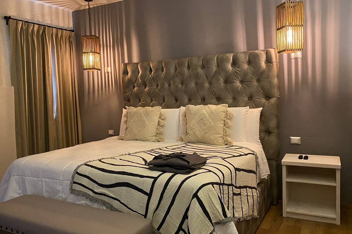 A bedroom with a tufted headboard, a bed with striped bedding, a bench at the foot of the bed, and two wall-mounted lamps.