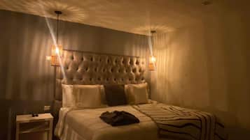 Un dormitorio con poca luz, un cabecero capitonado, una mesita de noche y una cama con manta a rayas.