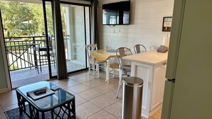 A modern kitchen with a dining area, a TV mounted on the wall, and a view of the outdoors through large sliding doors. - Castaways King Villa on Beach, Free Bikes! (Hilton Head Island)