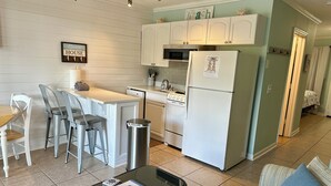 A compact kitchen with white cabinets, a refrigerator, and a stove. There is a dining area with a table and chairs, and a small seating area with a couch. - Castaways King Villa on Beach, Free Bikes! (Hilton Head Island)