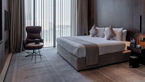 Een moderne hotelkamer met een groot bed, een zithoek met een stoel en tafel, en uitzicht op de stad door grote ramen.