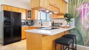 A modern kitchen with wooden cabinets, a black refrigerator, a built-in oven, a microwave, and a sink. There is a large window providing natural light and a decorative plant on the counter. - Steps to Beach | Private Balconies | Relax Here (Jacksonville Beach)