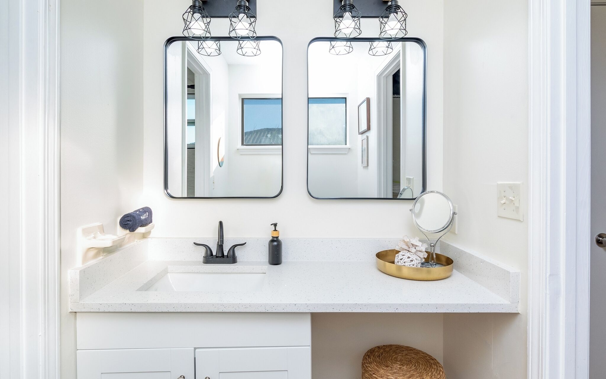 A bathroom with a white countertop, two mirrors, a sink, a soap dispenser, and a small round mirror on the wall.