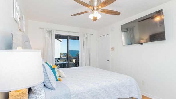 A bedroom with a bed, a ceiling fan, a large window with curtains, a TV mounted on the wall, and a balcony with a view. - Steps to Beach | Private Balconies | Relax Here (Jacksonville Beach)