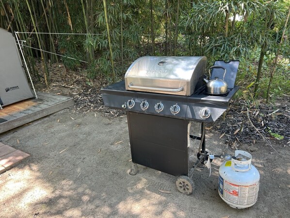 Una parrilla de gas portátil con cuatro quemadores e incluye ahumador, un tanque de propano y una pequeña tetera a un lado.