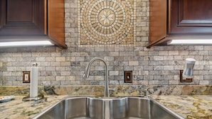 A kitchen with a double stainless steel sink, a granite countertop, a mosaic backsplash, and wooden cabinets.
