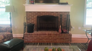 Una sala de estar con chimenea de ladrillo, un televisor de pantalla plana montado encima y una mesa de centro de vidrio con alcachofas.