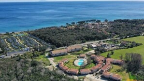 Luftaufnahme eines Küstenresorts mit mehreren bungalowsartigen Gebäuden, einem zentralen Swimmingpool und Blick auf Meer und Grünflächen.