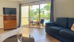 Un salon comprenant un canapé, un téléviseur, une table en verre avec une boisson, et une porte-fenêtre coulissante donnant sur un espace extérieur avec des chaises et une piscine.