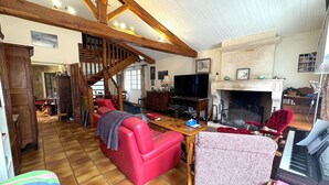 Un salon spacieux avec un canapé rouge, une table basse en bois et un piano. On aperçoit une cheminée, et un escalier mène à un autre niveau.