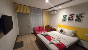 A hotel room with a bed, a red sofa, a small table, an air conditioner, and a TV mounted on the wall. - Kusma Hotel Bandungan Semarang (Bandungan)