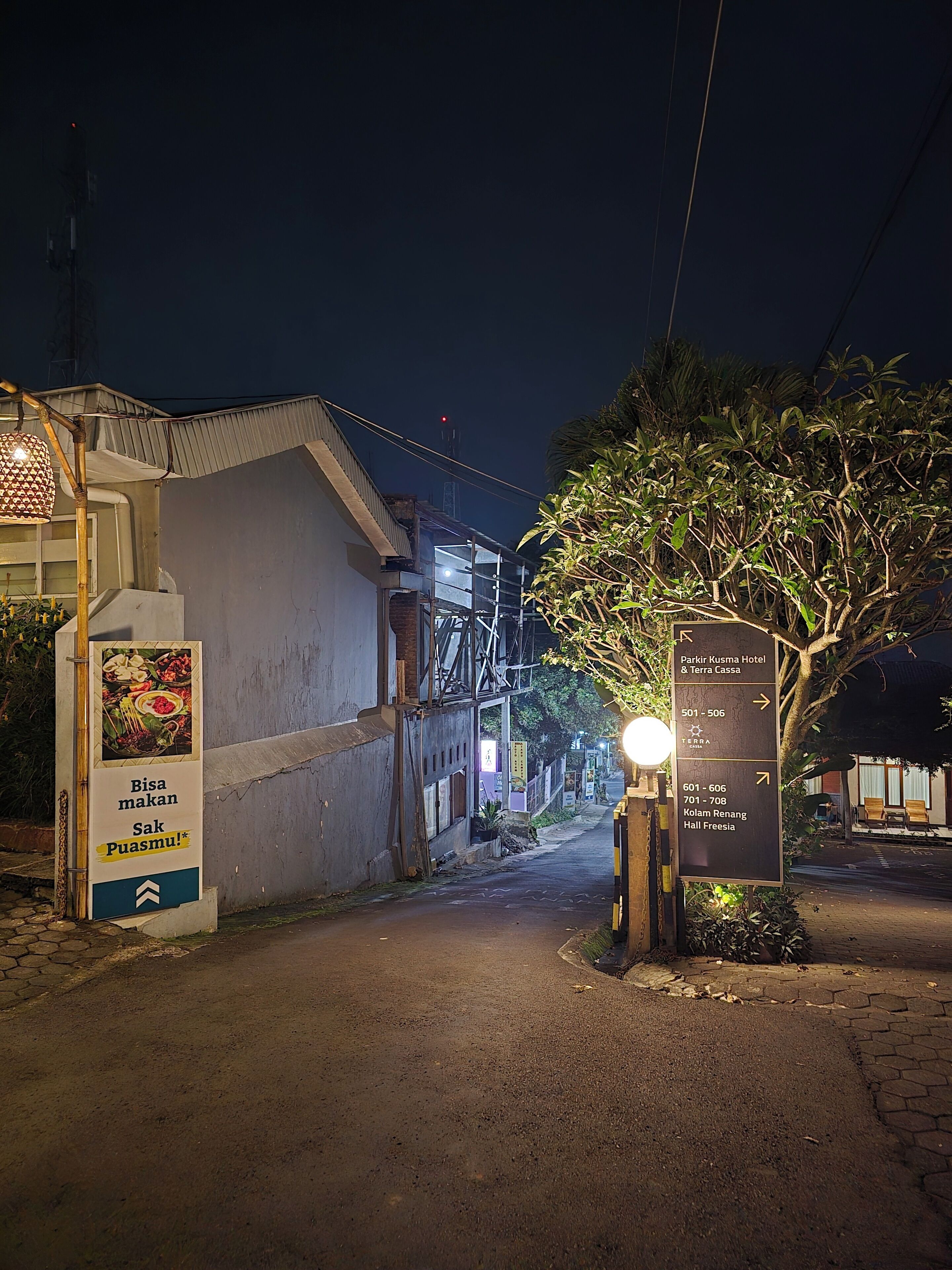 Escena nocturna de una calle con un letrero del Hotel Parkir Kusma & Terra Cassa, que indica las direcciones a diferentes habitaciones.