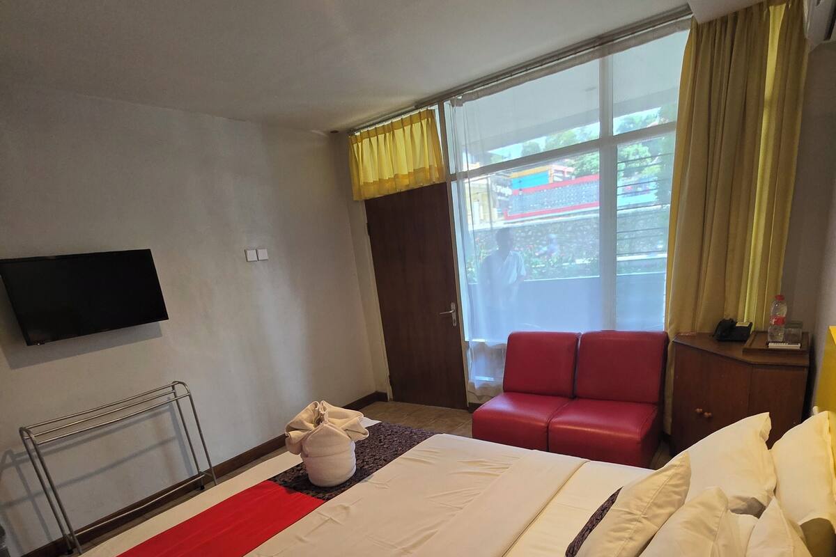 Ein Hotelzimmer mit einem großen Bett, einem roten Sessel, einem an der Wand befestigten Fernseher und einem Fenster mit Vorhängen.
