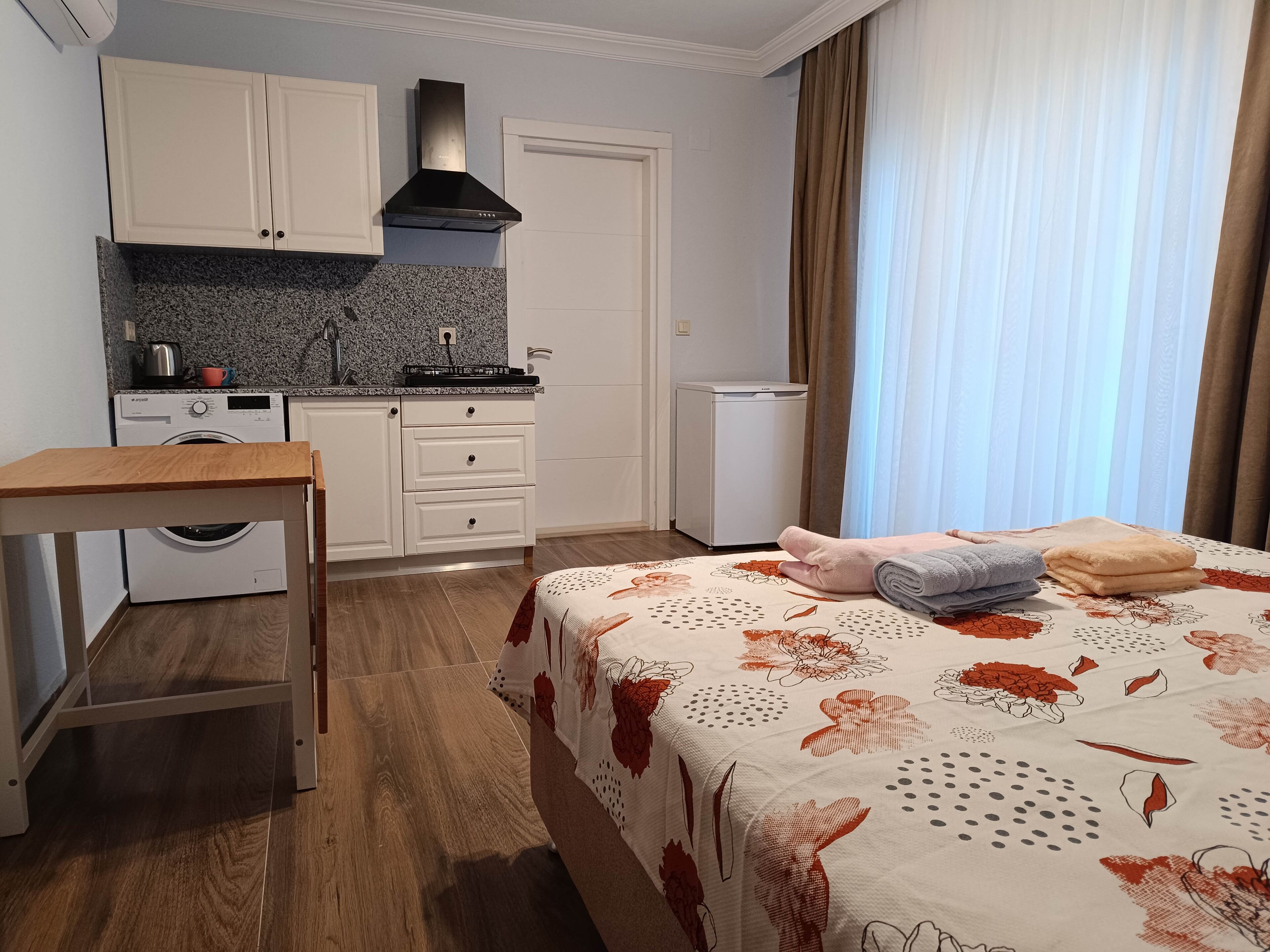 A compact hotel room with a kitchenette, a bed with floral bedding, a wooden table, and a washing machine.