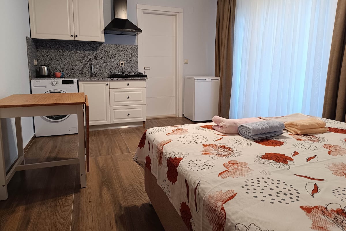 A compact hotel room with a kitchenette, a bed with floral bedding, a wooden table, and a washing machine.